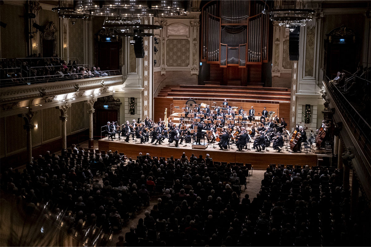 Ein Foto zeigt ein Sinfonieorchester bei einem Konzert. Die Musiker sitzen auf der Bühne und spielen ihre Instrumente.
